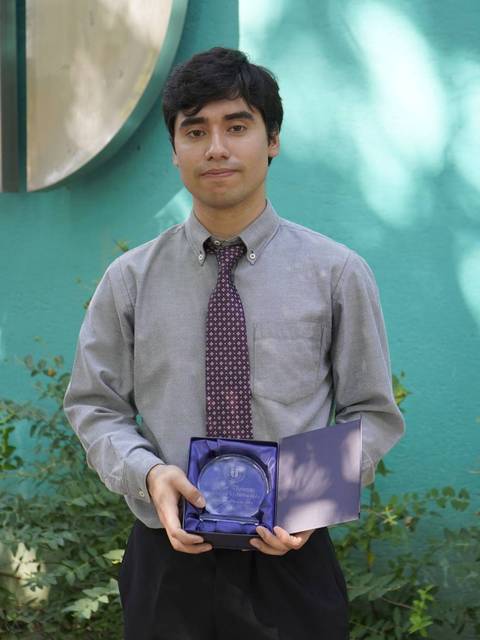 Un joven está posando con un premio en un entorno natural con un fondo colorido.
