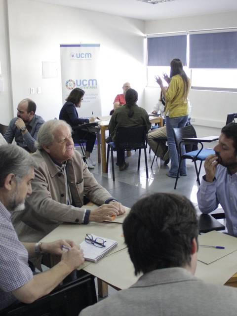 Un grupo de personas interactúa en una sala de reuniones, discutiendo en mesas de trabajo.