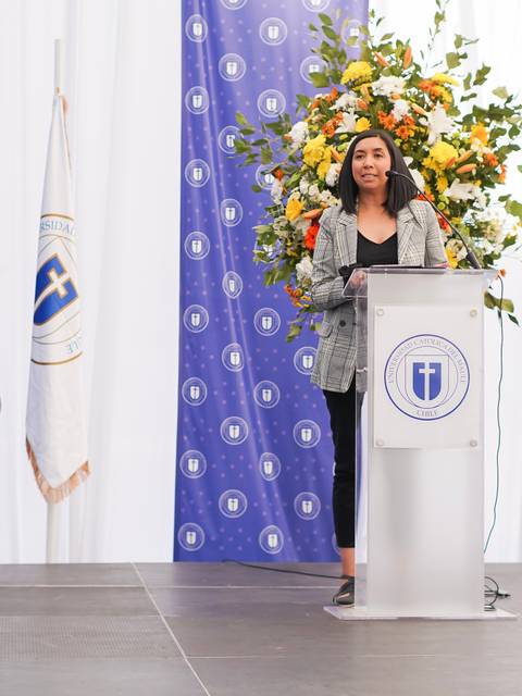 Una persona está de pie detrás de un podio en un evento formal, rodeada de decoración floral y banderas.