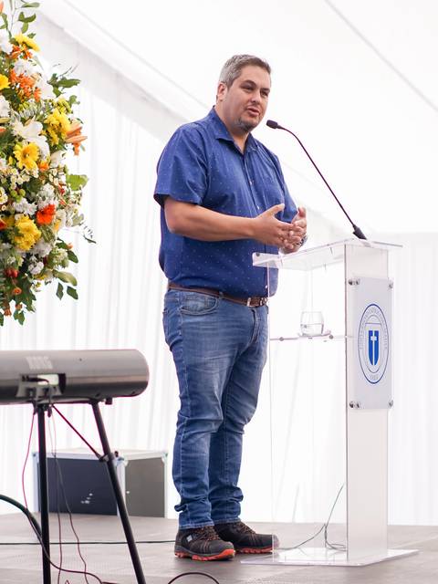Un hombre habla en un podio frente a un arreglo floral colorido en un evento al aire libre.
