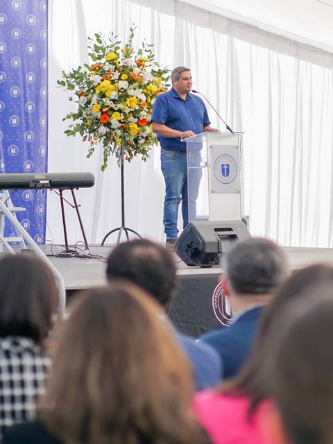 Un hombre habla en un podio durante un evento en un espacio cerrado decorado con flores.