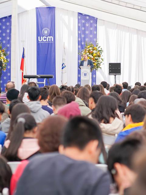 Un evento con una gran cantidad de personas sentadas en un auditorio decorado con flores y banderas.