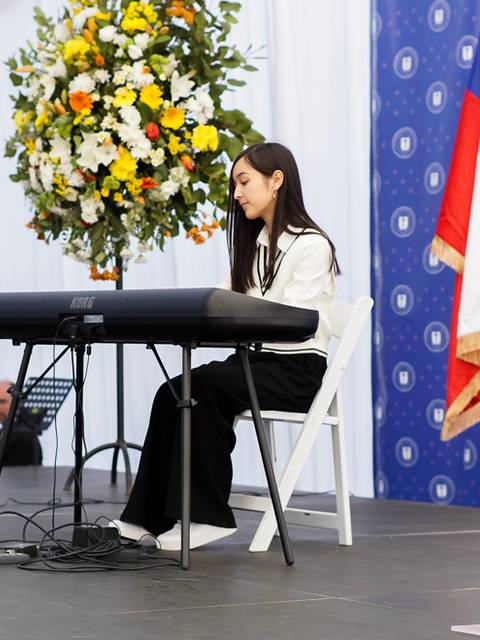 Una joven tocando un teclado frente a un auditorio decorado con flores y banderas.