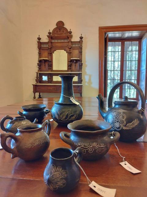 La imagen muestra una colección de utensilios de cerámica negra dispuestos sobre una mesa de madera, con un mueble decorativo al fondo.