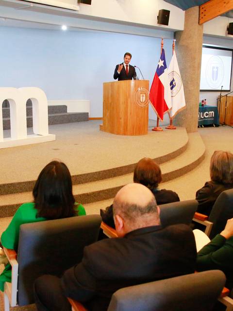 Un evento formal se lleva a cabo en un auditorio con un orador frente a una audiencia y decoraciones de fondo.