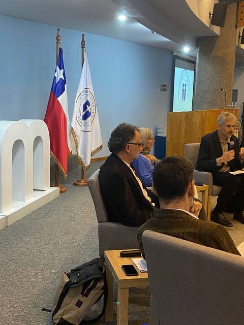 Un panel de discusión con varios oradores en un auditorio, decorado con banderas y el logotipo de una institución.