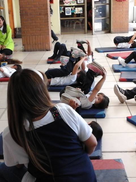 Un grupo de niños realiza ejercicios en el suelo de un aula mientras son supervisados por un adulto.