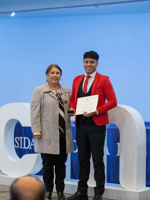 Una ceremonia de entrega de certificados donde un joven posa con una mujer frente a un fondo con el logo de la universidad.