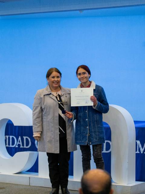 Dos mujeres posan junto a un letrero de la Universidad Católica del Maule mientras una de ellas sostiene un certificado.