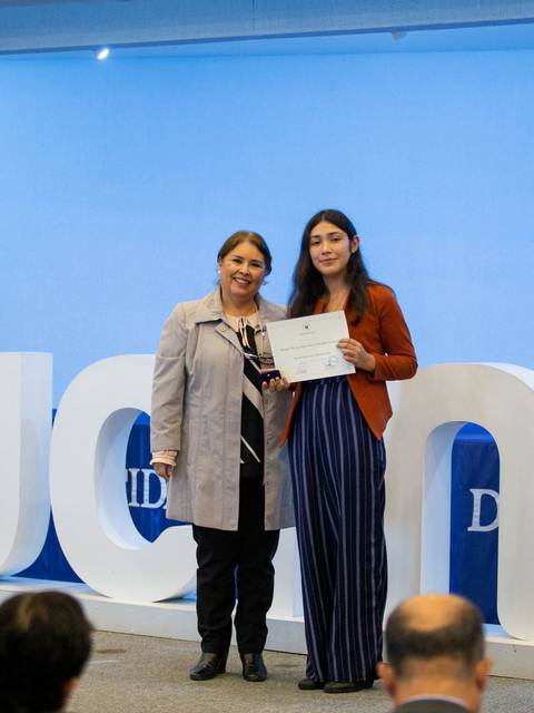 Una celebración de entrega de certificados en un evento con dos mujeres sonriendo, una de ellas sostiene un diploma.