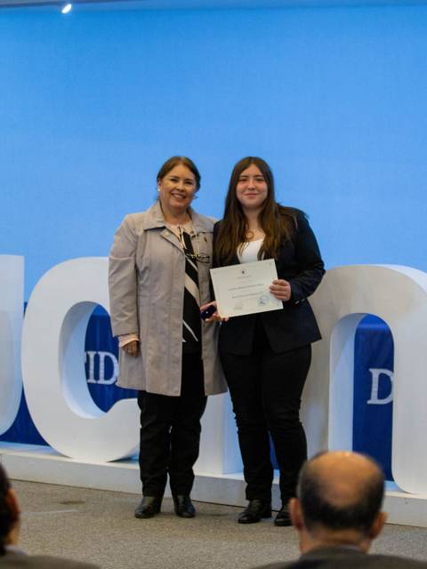 Dos mujeres posan en un evento de entrega de certificados junto a un fondo decorado.
