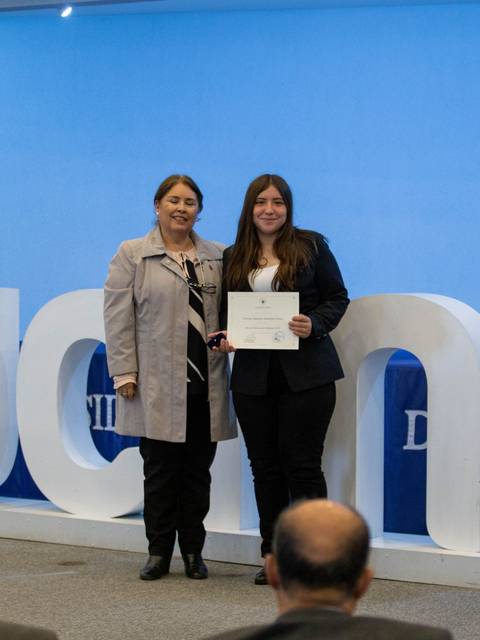 Una mujer entrega un certificado a una graduada en un evento formal.