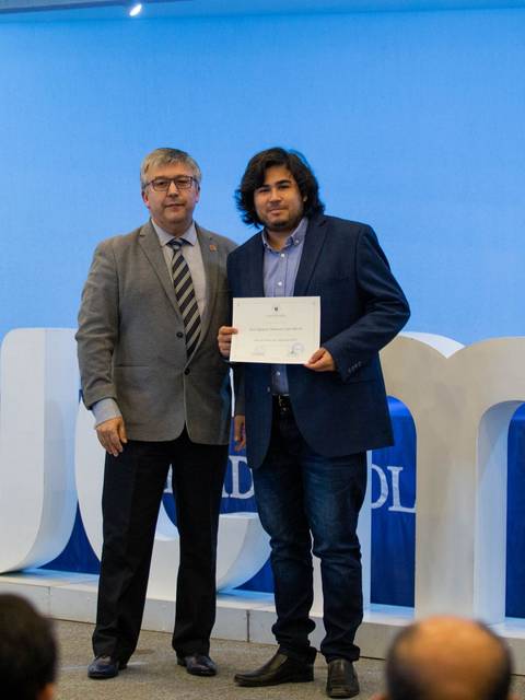 Dos hombres posan juntos en un evento de entrega de certificados en un ambiente formal.