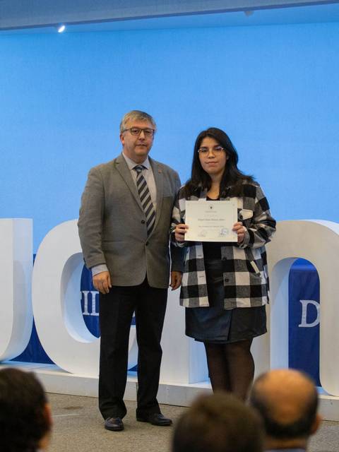 Una mujer recibe un certificado en un evento académico mientras un hombre la acompaña.