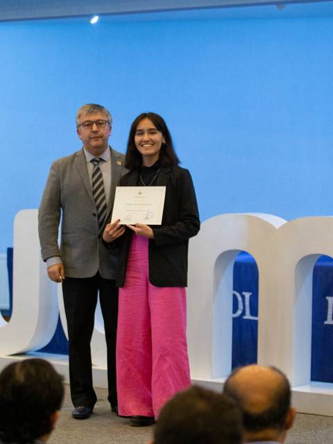 La imagen muestra a una joven recibiendo un certificado en un evento académico con decoraciones y un fondo institucional.