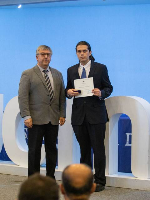 Una ceremonia de entrega de un certificado donde un hombre joven recibe un documento de un hombre mayor en un entorno decorado con flores.