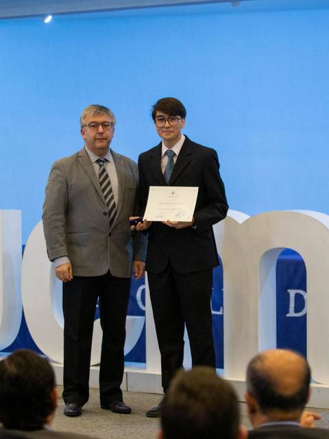 Un estudiante recibe un certificado en una ceremonia con un fondo decorativo y una cruz.