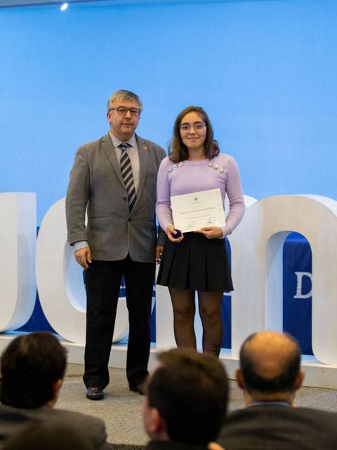Una estudiante recibe un diploma en una ceremonia formal en un auditorio decorado.