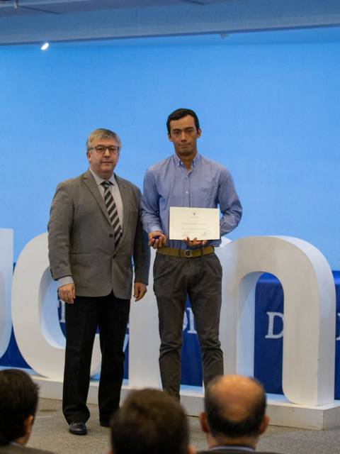 Dos hombres posan en un evento formal con un cartel de la Universidad de La Frontera y una cruz al fondo.