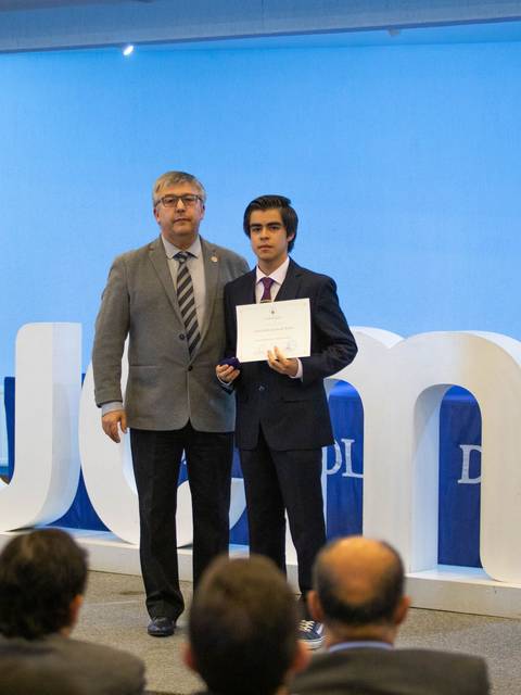 Un estudiante recibe un certificado en una ceremonia formal acompañada por un docente.