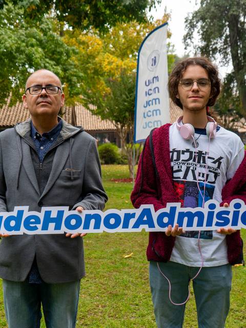Dos personas sostienen un cartel que dice '#BecaDeHonorAdmision' en un ambiente al aire libre.
