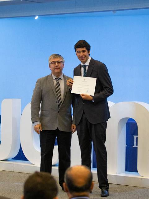 La imagen muestra a dos hombres posando en un evento de entrega de certificados, con un fondo decorado y una cruz visible.