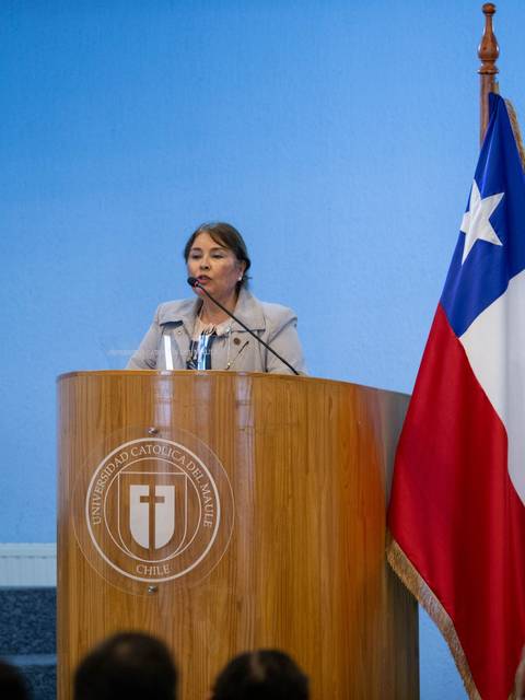 Una mujer habla desde un podio en un evento formal con banderas de Chile y de la institución detrás de ella.