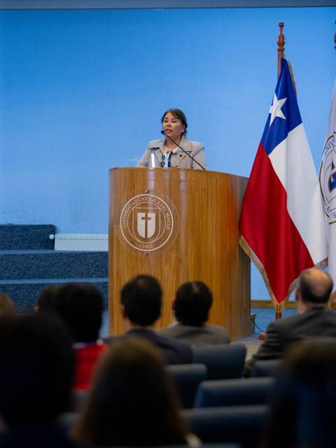 Una mujer habla desde un podio en un evento formal, con banderas de Chile y de una institución detrás de ella.