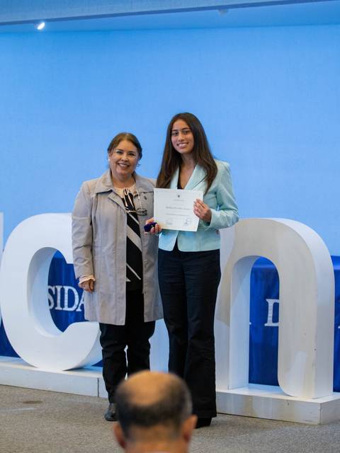 Dos mujeres posan en un evento, sosteniendo un certificado delante de un fondo con la sigla UCN.