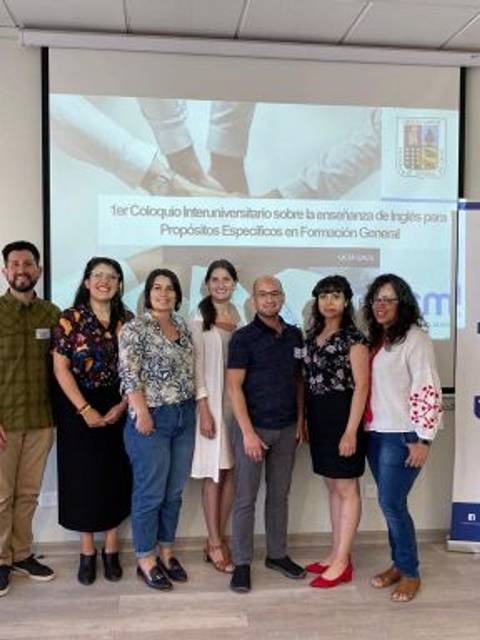 Un grupo de personas posando frente a una presentación sobre un coloquio interuniversitario.