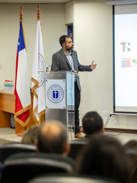 Un hombre presenta en un auditorio mientras se observan banderas chilenas al fondo.