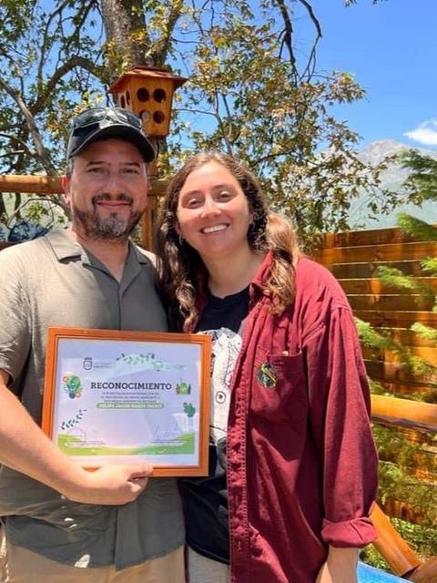 Una pareja sonriente sostiene un certificado de reconocimiento en un entorno natural.