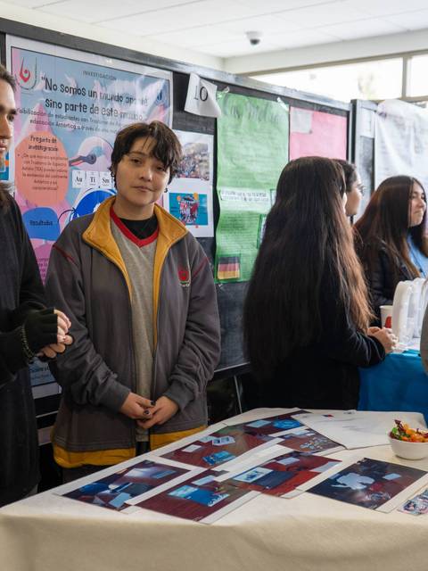 Estudiantes participan en una feria de proyectos escolares, mostrando sus trabajos y interactuando entre ellos.
