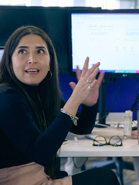 Una mujer con cabello largo y oscuro, vestida con un suéter negro, gesticula mientras habla en una oficina moderna.