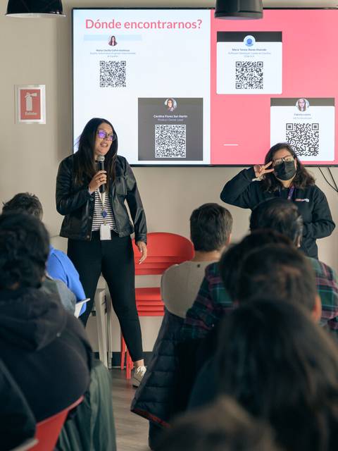 Un grupo de personas presenta información en una sala con pantallas mostrando códigos QR.