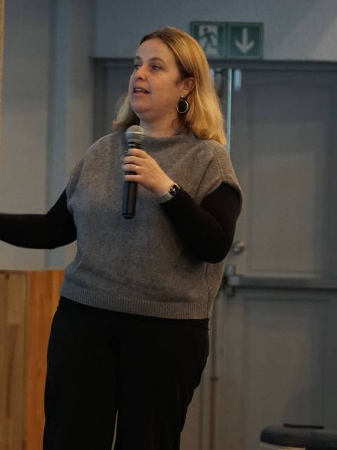 Una mujer presenta en un evento mientras un hombre la observa al fondo.