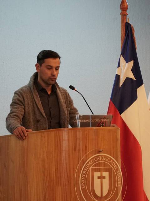 Un hombre habla en un podio frente a las banderas de Chile y de la Universidad Católica del Maule.