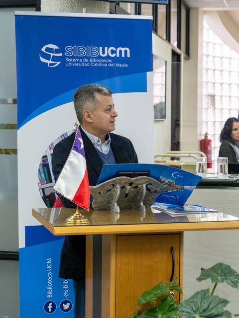 Un hombre presenta en un evento en una biblioteca universitaria con una bandera chilena y material promocional.