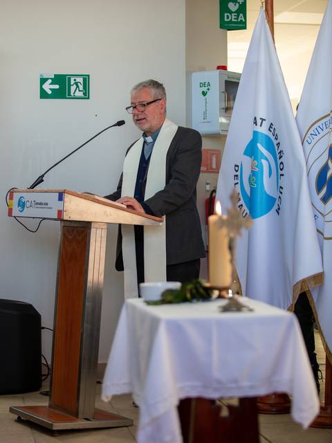 Un hombre vestido con traje y un alzacuellos da un discurso frente a un podio en un evento formal.