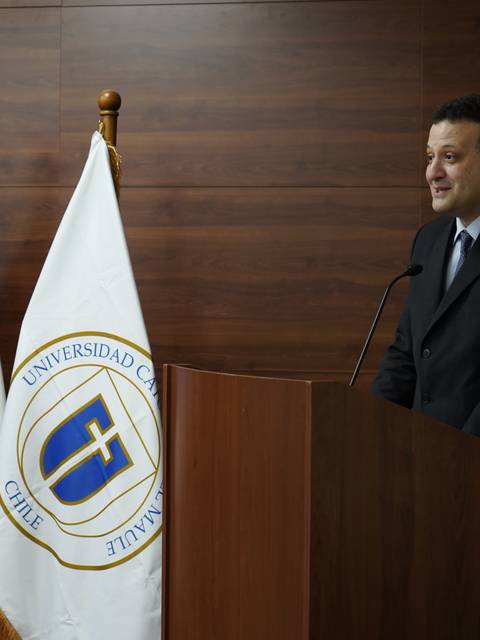 Un hombre está de pie frente a un podio, hablando en un evento formal con banderas chilena y de la universidad al fondo.