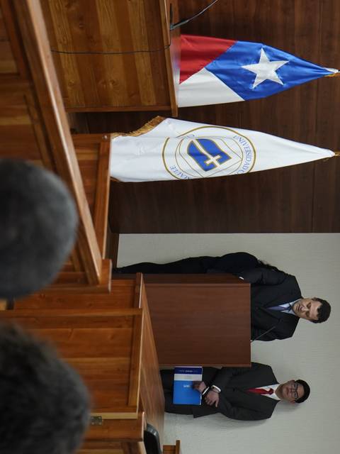 La imagen muestra a dos personas de pie en un recinto formal con banderas de Chile y una insignia en el fondo.