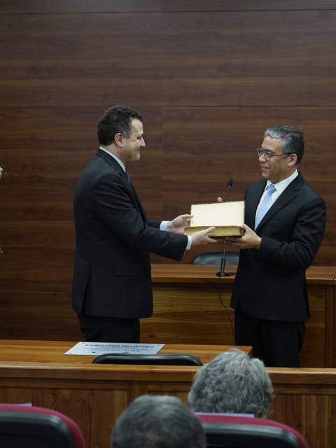 Dos hombres intercambian un obsequio en un evento formal rodeados de flores decorativas.