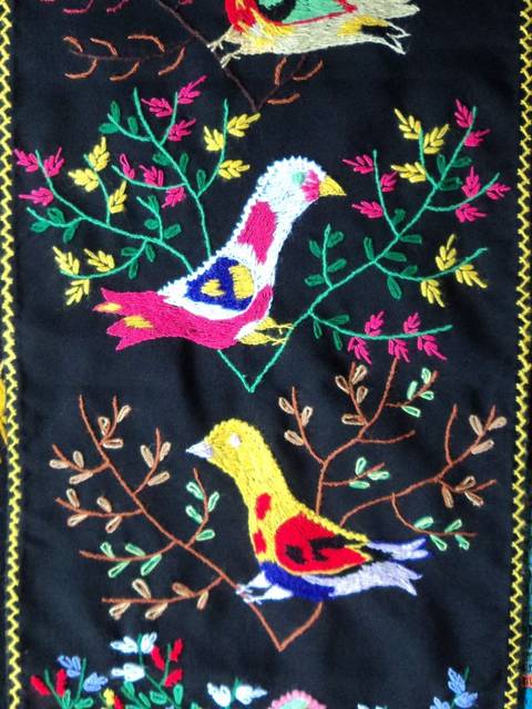 Una obra de arte textil que representa tres aves coloridas sobre un fondo negro, rodeadas de ramas y flores bordadas.