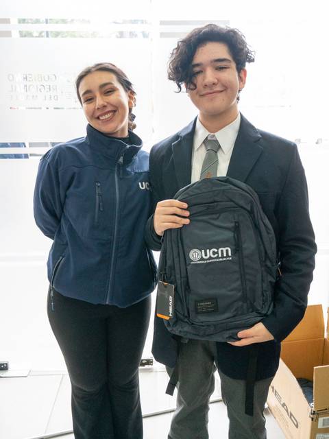 Dos jóvenes sonrientes posan juntos, uno de ellos sostiene una mochila con el logo de la UCM.