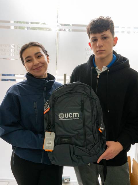Dos jóvenes posan juntos sosteniendo una mochila de color gris con el logo de UCM.
