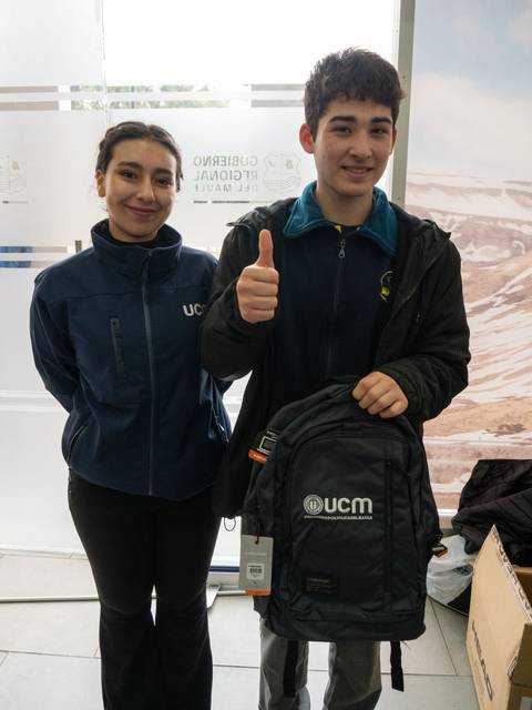 Dos jóvenes sonrientes posan juntos, uno de ellos muestra una mochila con el logo de UCM.