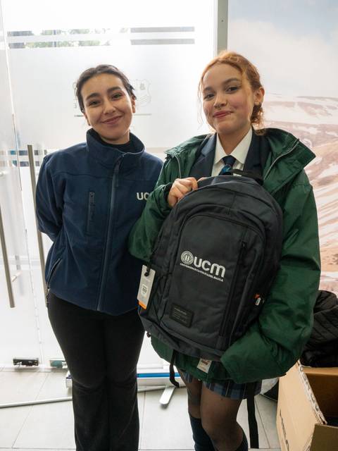 Dos personas sonrientes posan frente a una puerta sosteniendo una mochila con el logo de UCM.