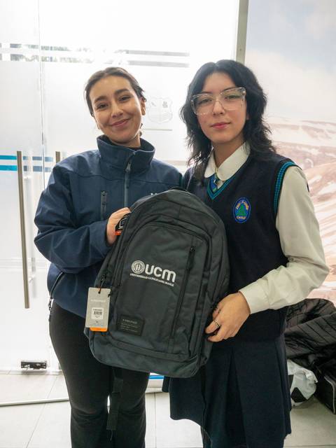 Dos mujeres posan sonrientes sosteniendo una mochila en un entorno interior.