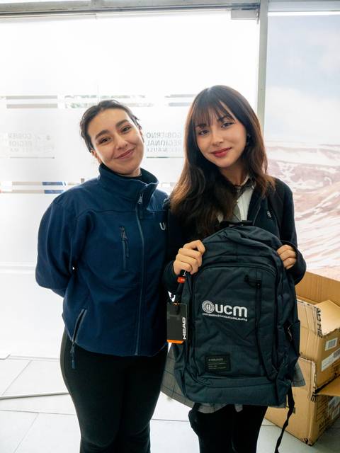 Dos mujeres sonríen mientras sostienen una mochila con el logo de UCM.