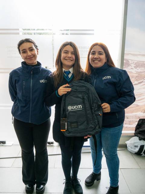Tres personas sonrientes posan juntas con una mochila en un ambiente interior.